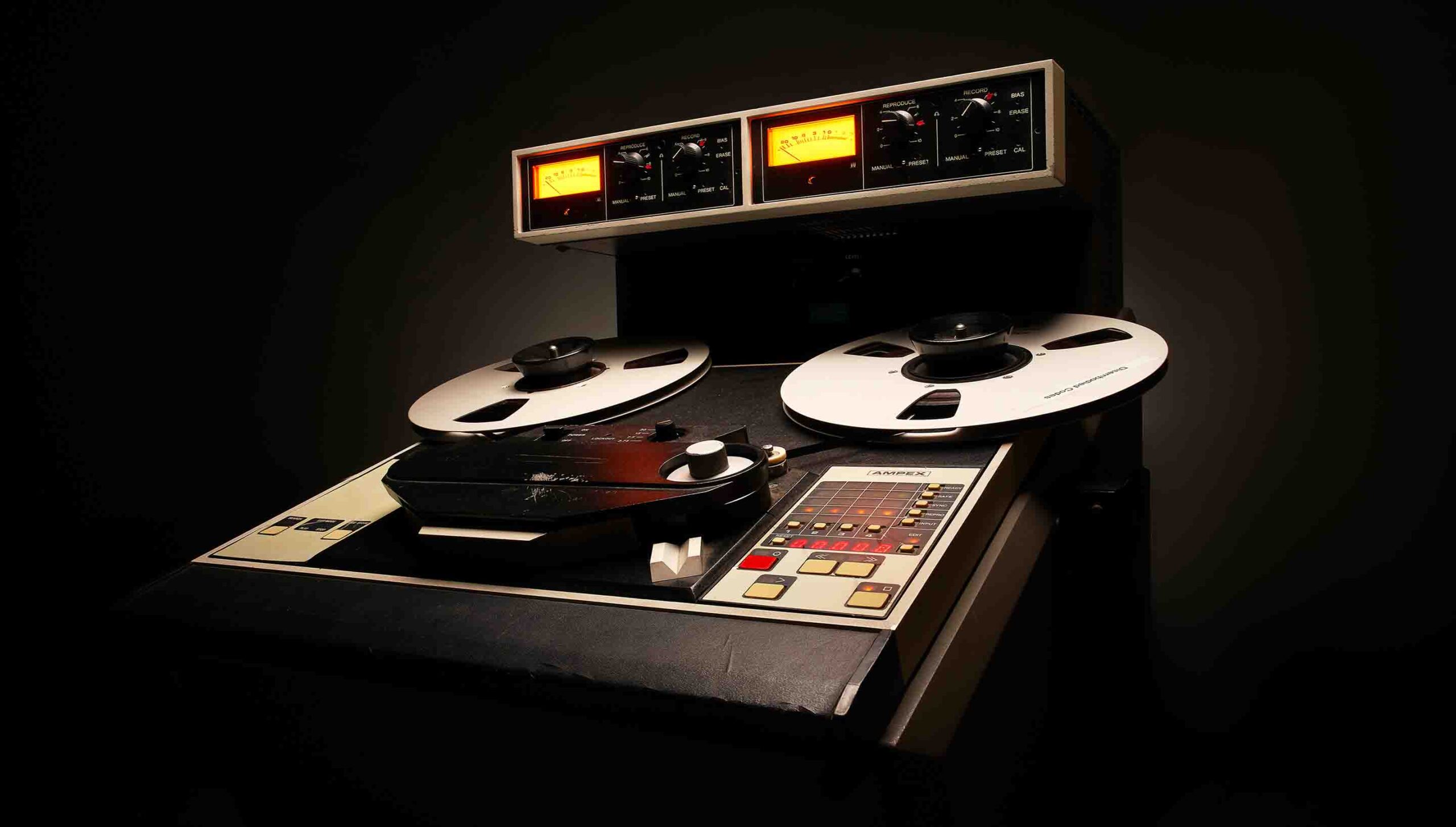 暗い部屋に置かれた Ampex ATR-102 の実機。上部にオレンジ色に光る2つのメーター、中央に大きなテープのリール（Universal Audio 公式の画像）